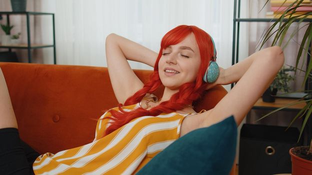 Happy young ginger girl wearing wireless headphones listening favorite energetic disco music song, relaxing, sleeping, resting, having a break, lying on couch at home. Weekends entertaining activities