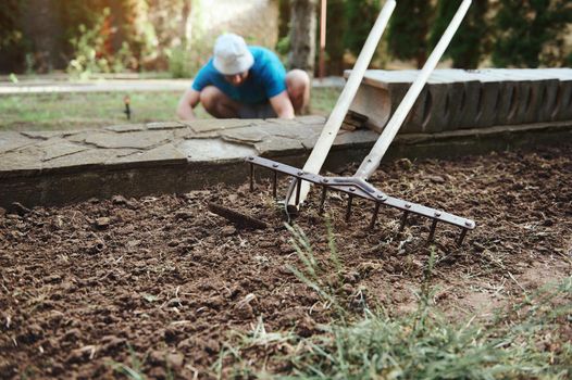Focus on a garden shovel and rake on the loose ground and farmer working on the field on blurred background. Agribusiness. New agricultural planting season. Agro Industry. Agriculture, organic farming