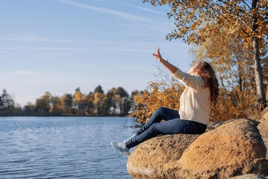 Freedom concept. Thoughtful romantic woman sitting on rocks on the river bank in sunset in autumn day, copy space.