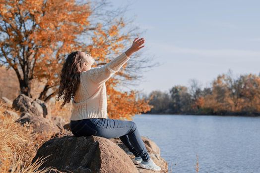 Freedom concept. Thoughtful romantic woman sitting on rocks on the river bank in sunset in autumn day, copy space.