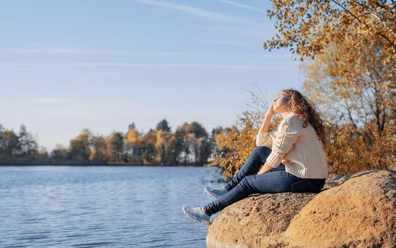 Freedom concept. Thoughtful romantic woman sitting on rocks on the river bank in sunset in autumn day, copy space.