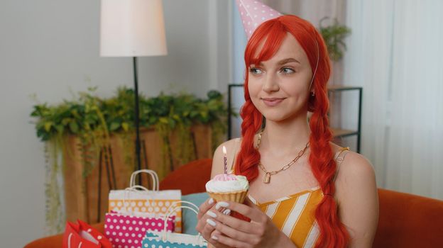 Happy red hair girl in festive birthday hat hold cupcake makes wish joyful congratulating blowing burning candle on cake. Young ginger woman celebrating anniversary party on couch with gifts at home