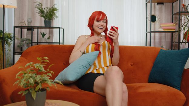 Excited happy redhead woman use mobile smartphone typing browsing say Wow yes found out great big win good news celebrate doing winner gesture. Young ginger girl at home in living room sitting on sofa