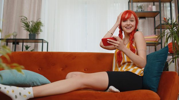 Worried funny young redhead woman enthusiastically playing shooter online video games on smartphone. Excited girl player using mobile phone gadget app celebrate win victory at home sitting on couch