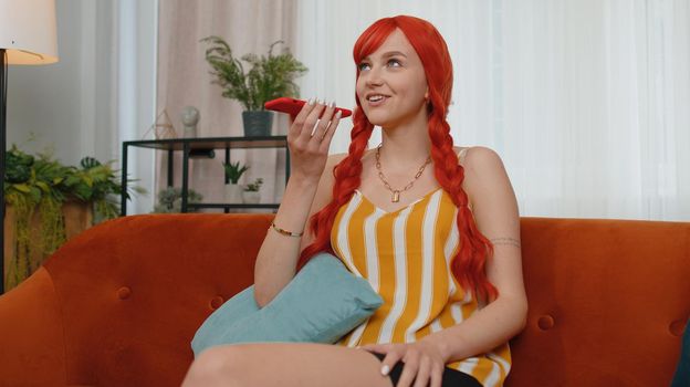 Smiling young redhead woman in tank top making phone conversation with friends sitting call on couch at home in room. Happy excited ginger businesswoman girl enjoying mobile loudspeaker talking indoor