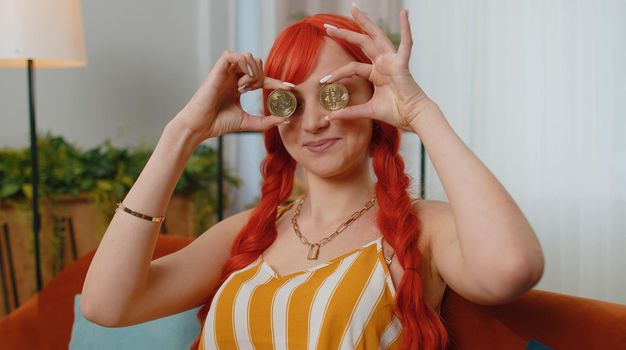 Woman freelancer holding two gold coins at home, successful developer programmer. Girl stock trader earning bitcoins after online monitoring trading operations. Increasing wealth, financial prosperity