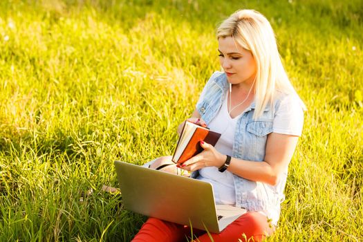 Young woman with laptop sits on the grass in the park on a sunny day. Laughing blonde in a white tank top and shorts. Online training, remote work and communication in social networks.