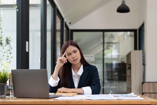 Asian woman sitting at desk near computer cogitating thinking making important decision at workplace. Concentrated serious office worker millennial woman analysing results feels doubts and feel unsure.