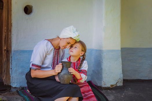 mother and daughter in Ukrainian national costumes are sitting near an old house