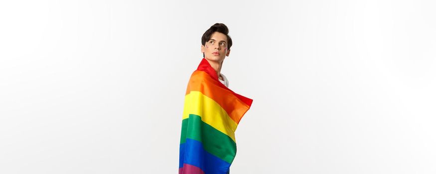 Image of thoughtful young gay man wearing lgbtq flag, turn head at upper left corner, staring at logo, standing over white background.