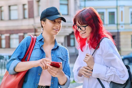 Two teenage females talking, looking into smartphone, outdoor on city street. Attractive girls friends, communicate together. Friendship, communication, adolescence, urban lifestyle, youth concept