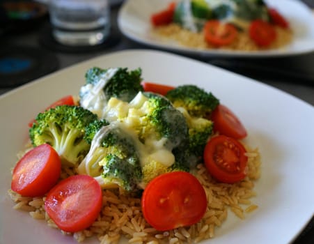 A delicious vegetarian meal with rice, small sliced ​​tomatoes, broccoli and melted cheese on a white plate. The same meal on another plate blurred in the background