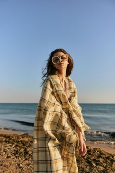 a woman wrapped in a plaid stands on the seashore in bright sunglasses against the background of the sunset sky. High quality photo