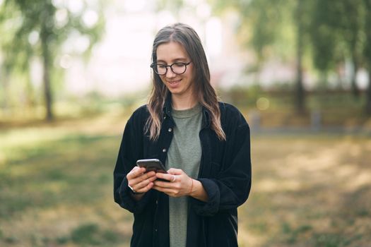 Young woman with glasses reading a message on her phone while standing in the park. High quality photo