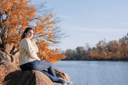 Freedom concept. Thoughtful romantic woman sitting on rocks on the river bank in sunset in autumn day, copy space.