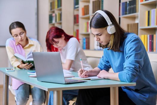 Group of teenage students study sitting at desk in library, in focus is guy in headphones using laptop book and writing in notebook. College, high school, education, learning, knowledge concept