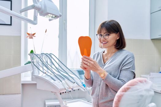 Happy middle aged woman dentist patient with a mirror in her hands looking at her teeth. Treatment, whitening, implants, medicine, health care and beauty smile concept