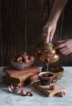 A woman pours tea from a copper teapot into a cup against the background of a stack of books and a cup with lychee, homely cozy tea drinking and reading,. High quality photo