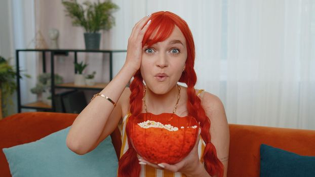 Excited young redhead woman sitting on sofa eating popcorn and watching interesting tv comedy serial, film, movie or social media content online at home. Ginger girl enjoying domestic entertainment