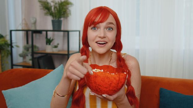POV of excited redhead woman sitting on sofa eating popcorn and watching interesting tv comedy serial, film, movie or social media content online at home. Ginger girl enjoying domestic entertainment