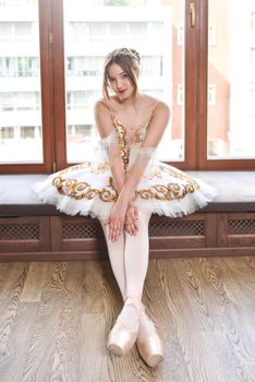 A pretty young ballerina in a white elegant tutu sits and poses on a window sill.