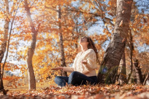 Autumn nature. Happy young woman throwing yellow leaves up in the air sitting in the autumn forest