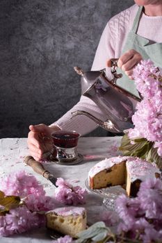 a woman pours tea from a silver teapot against a background of pink sakura flowers, a beautiful Easter cake stands on the table. High quality photo