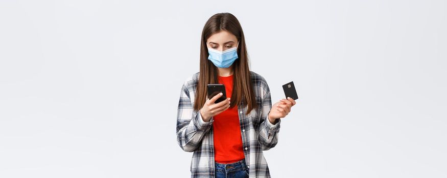 Coronavirus outbreak, working from home, online shopping and contactless payment concept. Girl in medical mask holding credit card, order clothes using mobile phone application.