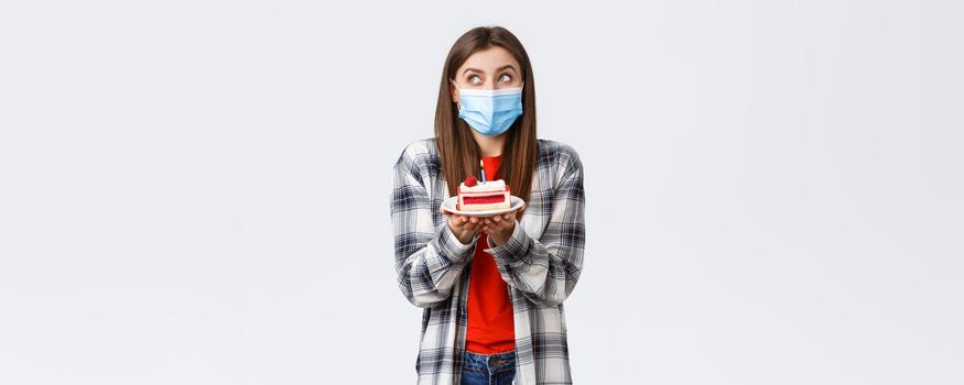 Coronavirus outbreak, lifestyle during social distancing and holidays celebration concept. Dreamy cute girl in medical mask, dreamy look up, imaging making wish as holding birthday cake with candle.