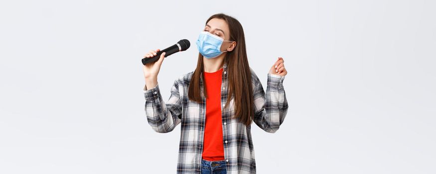 Coronavirus outbreak, leisure on quarantine, social distancing and emotions concept. Carefree young girl in medical mask, singing with microphone, close eyes, enjoying karaoke.