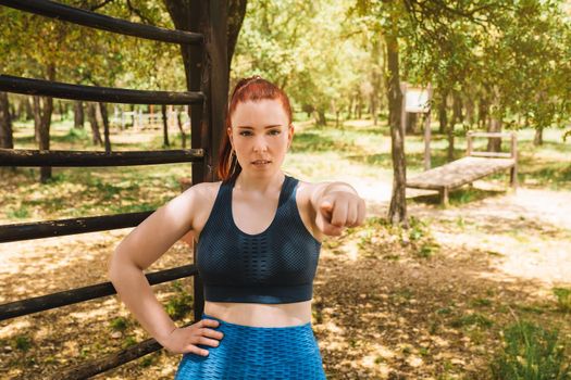 young female athlete pointing her index finger at the camera outside. young girl athlete challenging the viewer. health and wellness lifestyle. outdoor public park, natural sunlight.