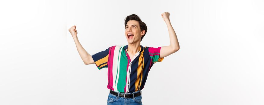 Happy gay man celebrating victory, raising hands up and rejoicing of winning, tirumphing while standing over white background.