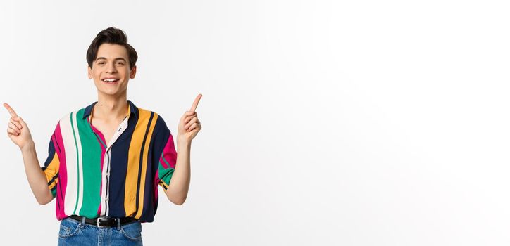 Happy young gay man pointing fingers sideways, showing two choices and smiling, standing against white background. Copy space