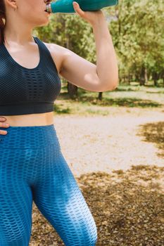 side image of a young fitness girl drinking water from a bottle after a sports workout to refresh herself. girl practising sport outdoors. health and wellness lifestyle.outdoor public park, natural sunlight. vertical