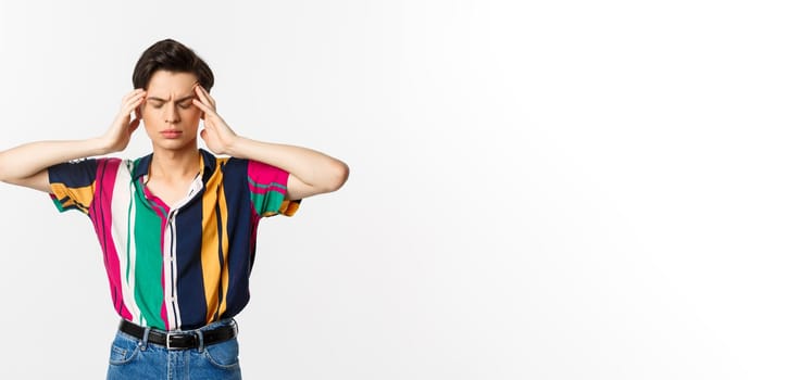 Image of young man having headache, looking troubled, holding hands on head, standing over white background.