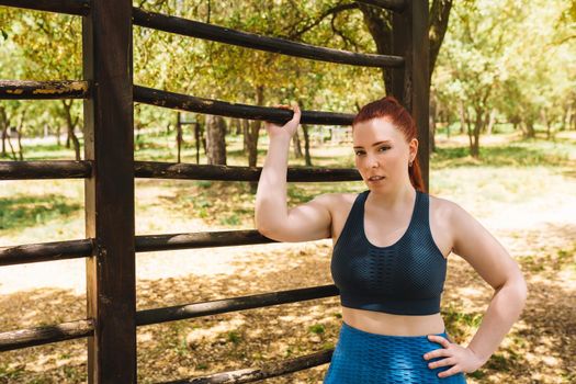 portrait of a young female athlete leaning on a public trellis outside. athlete practising sport outdoors resting after training. health and wellness lifestyle. outdoor public park, natural sunlight. vertical