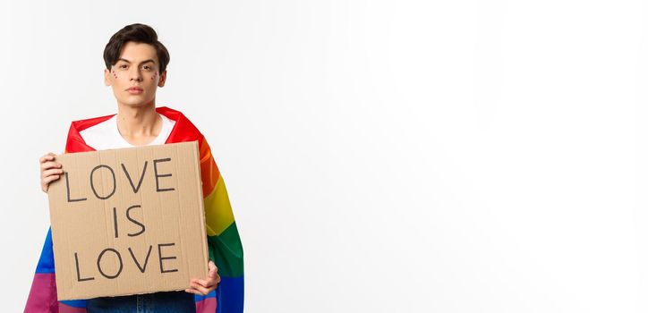 Serious and confident gay man wearing rainbow lgbt flag, holding sign for pride parade, standing over white background.