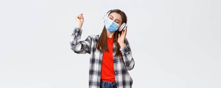 Social distancing, leisure and lifestyle on covid-19 outbreak, coronavirus concept. Carefree tender young woman carried away listening music in headphones, dancing with closed eyes in medical mask.