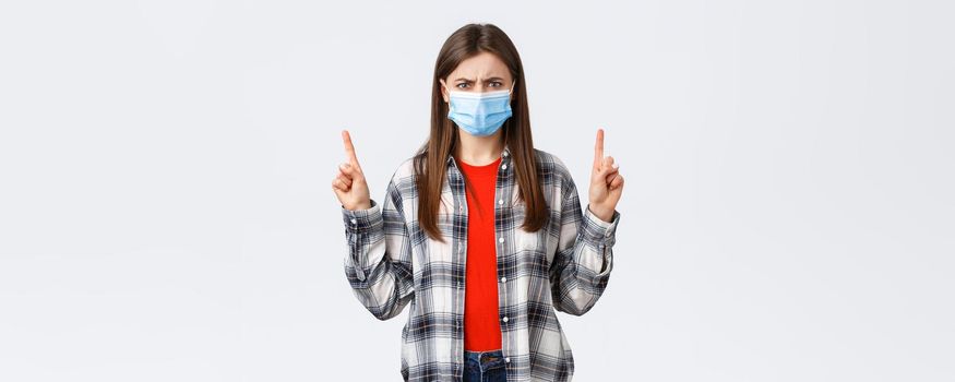 Coronavirus outbreak, leisure on quarantine, social distancing and emotions concept. Angry, outraged young woman in medical mask grimacing, looking mad as pointing up.