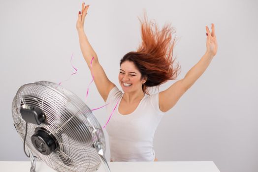 Joyful caucasian woman enjoying the wind blowing from an electric fan on a white background