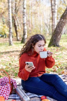 Leisure, free time. Beautiful caucasian woman in red sweater on a picnic outdoors, sitting on a plaid in autumn forest