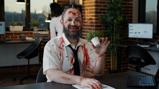 Nightmare zombie with bloody wounds smirking creepy while biting plant and waving at camera. Apocalyptic monster looking brain dead in office workspace while smiling mindless and evil.