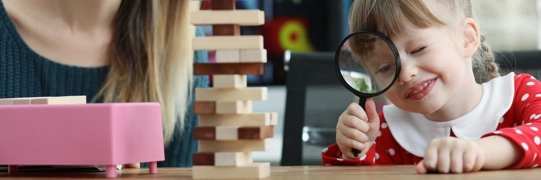 Portrait of cute toddler girl watch through magnifying glass on wooden tower. Teacher help child in development, improve concentration. Fun school concept