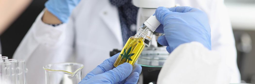 Close-up of persons hand hold jar with cannabis oil and going to add drop under microscope. Woman scientist explore sample, protective uniform. Lab concept