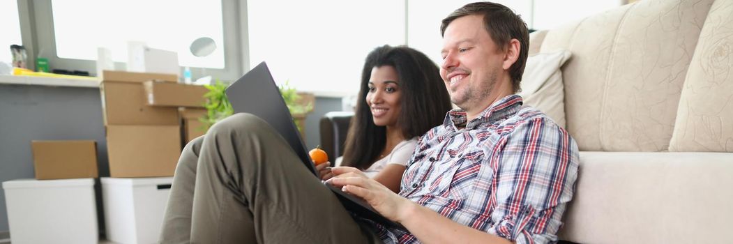 Low angle of cheerful smiling couple watch show online on laptop at home. Man and latin girl sit on floor, spend time on weekend. Relationship, fun concept