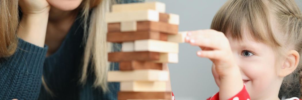 Portrait of beautiful little girl kid pull wooden block out of high tower and smile. Mum join daughter in playing board game. Parenthood, childhood concept