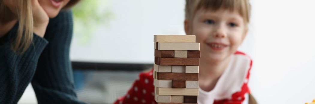 Portrait of happy mother with child girl playing wooden blocks tower at home. Stack and crash board game for fun. Family, pastime, entertainment concept