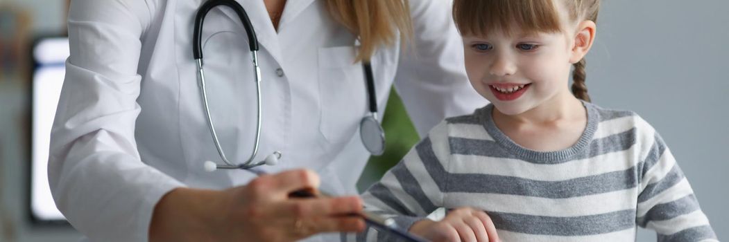 Portrait of pediatrician female try to distract child attention and make diagnostic. Smiling kid on planned checkup in clinic. Medicine, healthcare concept