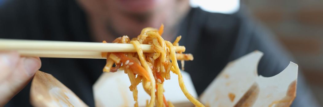 Close-up of smiling man eating spicy noodle chinese food with vegetable using chopsticks. Happy guy enjoy taste of takeaway portion. Hunger, lunch concept