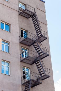 Silhouette of a fire escape on a high-rise building against a blue sky with clouds. Some of the stairs are broken. There is free space for text